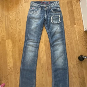 Blå jeansbyxor från Castina Denim med slitningar - Säljer ett par blå jeansbyxor från Castina Denim med slitningar och två patchdetaljer. Byxorna har raka ben, normal passform och dekorativa nitar på bakfickorna. Perfekta för en avslappnad stil. Kom privat för fler mått och bilder 🤗