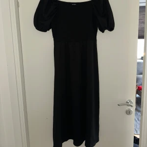 Svart långklänning med puffärm från Monki - Svart långklänning från Monki med smockad överdel och puffiga kortärmar. Klänningen har fyrkantig ringning och är tillverkad i ett mjukt, stretchigt material. Perfekt för dig som gillar en stilren och trendig look.