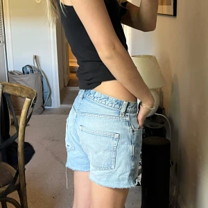 Ljusblå jeansshorts från Levi's - Säljer ett par klassiska ljusblå jeansshorts från Levi's med råa kanter och fem fickor. Shortsen har normal passform och är högmidjade. Perfekta för varma dagar och enkel att matcha med olika toppar.