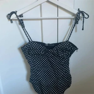 Svart body med vita prickar från Zara - Jätte populär body från Zara säljer för bra bud!!! storlek M men sitter som S-xs .Supersnygg svart body från Zara med vita prickar och knytbara axelband. Klassisk och trendig modell som ger en retrovibe. Perfekt för stranden eller poolen. Materialet är mjukt och stretchigt syntet. 