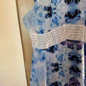 Blå blommig klänning med spetsdetalj strl: L men är mer som M - Säljer en blå och vit blommig klänning med smala axelband och bred vit spetsdetalj i midjan. Klänningen har en lätt och luftig känsla, perfekt för varma dagar. Mönstret är i olika blå nyanser och ger en fräsch look.