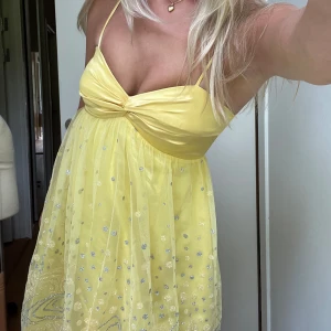 Gul klänning med paljettdetaljer - Söt gul klänning med smala axelband och knutdetalj framtill. Klänningen har glittriga paljetter och ett lätt, luftigt tyg som faller fint. Perfekt för sommarens alla tillfällen.