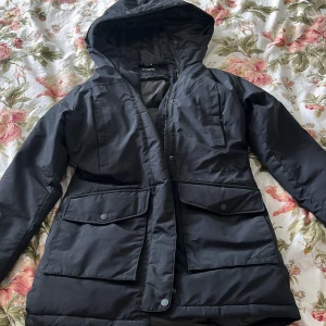 Svart vinterjacka med huva från Everest - Säljer en svart varm vinterjacka från Everest med huva och stora fickor framtill. Jackan har dragkedja och knappar, samt en klassisk parkamodell som passar perfekt för kalla dagar. Praktisk och stilren design. Används pga ingen användning 