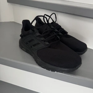 Svarta Adidas sneakers - Säljer ett par helsvarta Adidas sneakers med klassiska tre ränder på sidan. Skorna har snörning och är tillverkade i syntetmaterial och mesh för bra andningsförmåga. Perfekta för vardagsbruk eller träning.
