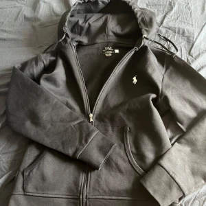 Svart hoodie från Polo Ralph Lauren - Svart hoodie från Polo Ralph Lauren med dragkedja och vit broderad logga på bröstet. Tröjan har huva med snörning och två fickor framtill. Perfekt för en avslappnad och stilren look.