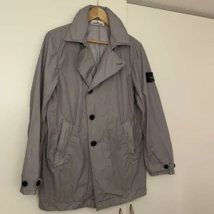 Ljusgrå trenchcoat från Stone Island - Säljer en ljusgrå trenchcoat från Stone Island med klassisk krage och svarta knappar. Jackan har två snedställda fickor framtill och ikonisk logotyp-patch på ärmen. Tillverkad i lätt syntetmaterial (Naslan Light Watro) som är slitstarkt och vattenavvisande.