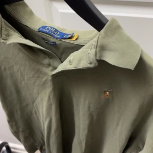 Olivgrön pikétröja från Polo Ralph Lauren - Säljer en klassisk olivgrön pikétröja från Polo Ralph Lauren med broderad logga på bröstet. Tröjan har krage, knappar vid halsen och korta ärmar. Perfekt för en stilren och avslappnad look.