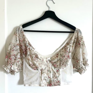 Blommig croppad korsettopp med puffärmar - Superfin blommig korsettopp!  Toppen är croppad och har en som en somrig vintagekänsla med sitt tunna, luftiga material och v-ringning tillsammans med den söta blommiga detaljerna. 