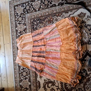 Randig långkjol i pastellfärger - Säljer en långkjol med volanger i mjuka pastellfärger som orange, rosa och beige. Kjolen har ett randigt mönster och är luftig med flera lager. Perfekt för dig som gillar boho-stil och vill ha något färgglatt till sommaren.