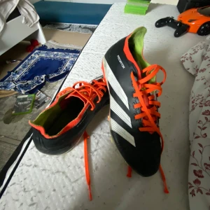 Adidas Predator fotbollsskor med orange snörning - Säljer ett par Adidas Predator fotbollsskor i svart med vita ränder och orange snörning. Skorna har dobbar för spel på gräs och en grön insida. Perfekta för dig som vill sticka ut på planen!