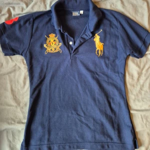 Marinblå pikétröja från Polo Ralph Lauren - Mörkblå pikétröja från Polo Ralph Lauren med broderade detaljer på bröstet och röd siffra på ärmen. Klassisk krage och korta ärmar. Perfekt för en sportig och stilren look. Inte så stor, utan passar mer XS till M. 