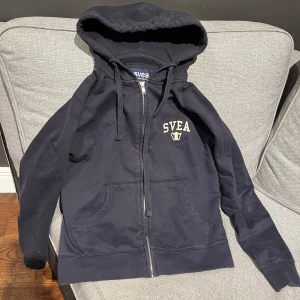 Mörkblå hoodie med dragkedja från Svea - Mörkblå hoodie från Svea med dragkedja framtill och två fickor. Svea-logga i vitt på bröstet. Klassisk huva och ribbade muddar vid ärmslut och nederkant. Perfekt för en avslappnad stil.