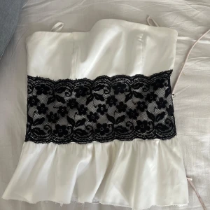 Vit tubtopp med svart spets från Vero Moda - Säljer en vit tubtopp från Vero Moda i satinliknande material med bred svart spetsdetalj över midjan. Toppen har volang nertill och tunna axelband. Perfekt för dig som vill ha något unikt och snyggt!