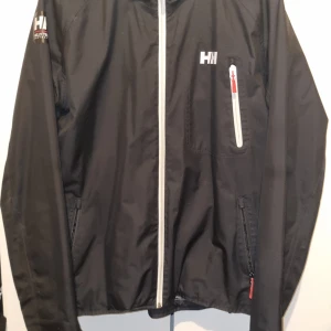 Svart vindjacka från Helly Hansen - Svart vindjacka från Helly Hansen med vit dragkedja framtill och på bröstficka. Jackan har två sidofickor med dragkedja och logga på bröstet samt ärmen. Perfekt för blåsiga dagar.