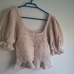 Beige stickad croppad topp med volang - Supersöt beige stickad topp med puffärmar och volangkant nertill. Den har ett vackert mönster och en djup rundad ringning. Perfekt för dig som gillar romantisk och bohemisk stil.