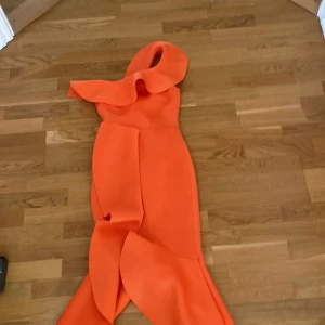 Orange oneshoulder klänning från True Violet - Säljer en orange statementklänning från True Violet med asymmetrisk oneshoulder och volangdetaljer. Klänningen har figurnära passform och är tillverkad i ett kraftigt, strukturerat material. Perfekt för dig som vill sticka ut. Använd endast en gång. 