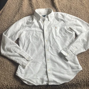 Vit skjorta från Ralph Lauren - Klassisk vit skjorta från Ralph Lauren med broderad logga på bröstet. Skjortan har lång ärm, knappar framtill och vid manschetterna samt button-down krage. Perfekt för en stilren och clean look.
