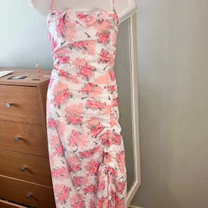 Blommig långklänning med slits - Säljer en romantisk långklänning med blommigt mönster i rosa och ljusa toner. Klänningen har smala axelband, en lätt draperad urringning och en slits framtill för extra rörelsefrihet. Perfekt för sommarens tillfällen!