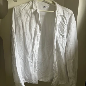 Vit randig skjorta från Zara - Vit skjorta från Zara med tunna vertikala ränder i ljusblått. Klassisk krage, långärmad och med bröstficka samt knappar framtill. Skjortan har en luftig känsla och passar perfekt till en avslappnad stil.