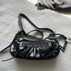Svart axelväska från Rizzo - Snygg svart axelväska från Rizzo i glansigt skinn med silverfärgade nitar och dragkedja framtill. Väskan har en cool, böjd form och detaljerade sömmar. Perfekt för att lyfta din outfit med en edgy touch. Väskan är helt ny och oanvänd! 