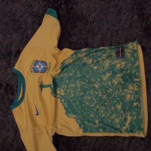 Brasilien Neymar 10 fotbollströja - Gul Brasilien fotbollströja med gröna detaljer, Neymar och nummer 10 på ryggen. Tryckt landslagsemblem på bröstet och Nike-logga. Unikt grönt mönster på framsidan. Perfekt för fans av landslaget.