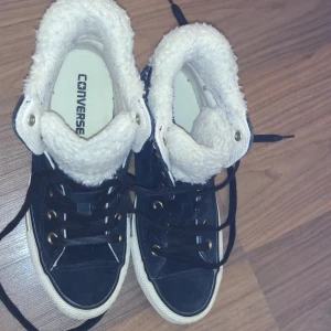 Svarta Converse sneakers med foder - Säljer ett par svarta Converse sneakers med högt skaft och vit sula. Skorna har mysigt vitt foder på insidan och guldiga öljetter för snörningen. Perfekta för kyligare dagar när du vill ha både stil och värme.