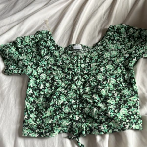 Grön blommig croppad blus - Säljer en söt blommig blus i grönt och svart med korta ärmar och knyt framtill. Toppen har en croppad passform och ett lätt rynkat tyg som ger en fin siluett. Perfekt för vår och sommar!  Den är lite liten i storlek 