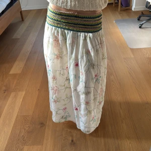 Blommig långkjol från ZARA - Superfin långkjol från ZARA med broderade blommor i rosa, grönt och gult på vit botten. Kjolen har en bred, elastisk midja med virkade detaljer i grönt, gult och rosa. Perfekt för en somrig och avslappnad look.