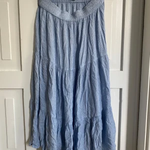 Ljusblå långkjol från Magasin du Nord - Säljer en ljusblå långkjol från Magasin du Nord. Kjolen har en avslappnad passform med resår i midjan och flera volanger som ger ett flowy intryck. Perfekt för varma dagar och enkel att styla med en enkel topp.