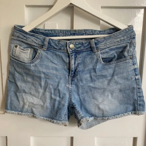 Ljusblå jeansshorts med låg midja - Säljer ett par ljusblå jeansshorts med låg midja. Klassisk femficksmodell med råa kanter nertill och bälteshällor. Perfekta för varma dagar och enkel att matcha med olika toppar.