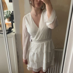 Vit omlottklänning med volanger - Superfin vit kortklänning med omlott-topp, långa transparenta ärmar och volangdetaljer i kjolen. Klänningen har markerad midja och lätt v-ringning. Perfekt för dig som tar studenten eller dig som bara vill ha en fin sommarklänning!           Klänningen är aldrig använd då jag valt en annan klänning till min student! Köpt på bubbleroom för 699kr!