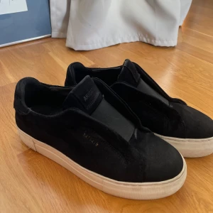 Svarta sneakers från ARIGATO - Säljer ett par svarta sneakers från ARIGATO med vit sula. Skorna har en stilren design utan snörning och är tillverkade i mocka. Perfekta för dig som gillar minimalistisk stil och vill ha ett par bekväma vardagsskor. Kartongen medkommer om man vill. Pris går att diskutera
