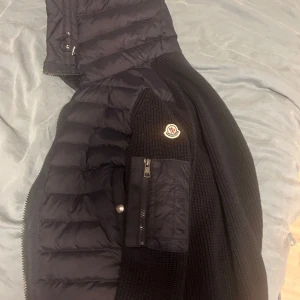 Moncler cardigan  - Mörkblå pufferjacka från Moncler med stickade ärmar och vadderad kropp. Jackan har en huva, dragkedja framtill och en praktisk ficka med dragkedja på ärmen. Ikoniska Moncler-loggan sitter på vänster ärm. Perfekt för kyliga dagar.