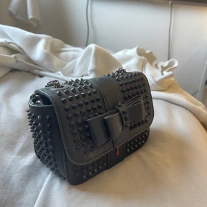 Svart axel väska -  Svart axelväska i svart skinn med coola nitar över hela väskan och en stor rosett framtill. Kedjerem i metall och röd insida som ger extra edge. Perfekt för dig som vill sticka ut med en statementväska.