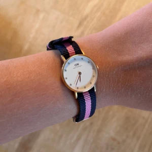 Daniel Wellington klocka med extra band - Daniel Wellingtonklocka i guld. Använd några få gånger bara. Medföljer tre utbytbara band: ett randigt i marinblått och rosa, ett randigt i marinblått och vitt samt ett brunt läderband. Kom med bud :-)