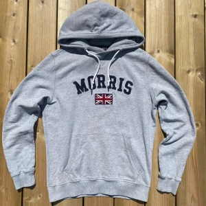 Grå hoodie från Morris med flagga - Snygg ljusgrå hoodie från Morris med broderad Union Jack-flagga och mörkblå text på bröstet. Tröjan har huva med snörning, känguruficka och ribbade muddar. Perfekt för chill dagar och streetstyle. Har några defekter som visas på bild 3 och 4. Metallen på snörena är även tillbucklade. Storlek Small, slim fit skulle jag säga. Vid frågor eller funderingar skicka ett meddelande💫