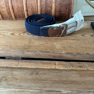 Mörkblå flätat bälte med skinn-detaljer - Snyggt mörkblått bälte med flätad design och bruna skinn-detaljer vid spännet. Bältet har ett klassiskt silverfärgat spänne och passar perfekt till jeans eller chinos. Materialet är mjukt och flexibelt för extra komfort.