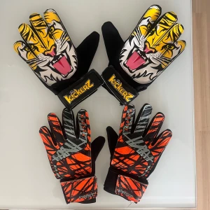 Två par målvaktshandskar från Kickerz och Kipsta - Säljer två par coola målvaktshandskar. Säljer både separat och i paket! Ett par från Kickerz med tigertryck i gult, svart och vitt, och ett par från Kipsta i orange och svart med grå detaljer. Båda har breda kardborreband vid handleden för bra passform.