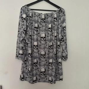 Grå klänning med dödskallemönster från SHEIN - Cool grå klänning från SHEIN med svartvitt dödskalle- och korsben-mönster. Klänningen har trekvartsärm och en lös passform, perfekt för dig som gillar edgy stil. Tyget är mjukt och lätt, vilket gör den bekväm att bära.
