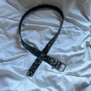 Svart skärp med punkmönster - Svart bälte med vita punkinspirerade tryck som säkerhetsnålar, anarkisymboler, texter som 'ANTI-SOCIAL' och 'ANARCHY' samt coola doodles. Skärpet har en silverfärgad metallspänne och är gjort i skinn. Perfekt för dig som gillar alternativ stil.