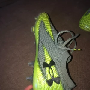 Under Armour grå/gula fotbollsskor - Snygga fotbollsskor från Under Armour mönstrad sula med fasta dobbar för bra grepp på planen. Perfekta för dig som vill sticka ut på matchen. Kolfibrer under som ger dig extra fjong när du ska springa.                                                                      Storlek 42,5       Nypris 2100.                                                Art:302828-348