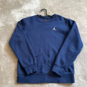 Blå crewneck från Jordan - Mörkblå sweatshirt från Jordan med klassisk Jumpman-logga på bröstet och broderad signatur på ärmen. Tröjan har rund halsringning och ribbade muddar vid ärmslut och nederkant. Perfekt för en avslappnad stil.