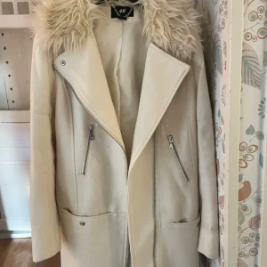 Beige kappa med fuskpäls från H&M - Snygg beige kappa från H&M med stor krage i fuskpäls. Jackan har dragkedjor framtill, två fickor med dragkedja och en rak, längre passform. Perfekt för dig som vill ha en stilren och trendig look under höst och vinter.