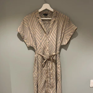 Beige-mönstrad midiklänning - Snygg beige midiklänning med mönster. Klänningen har korta ärmar, knytband i midjan och v-ringad hals. Lätt och flowy känsla, perfekt både vår, sommar och höst.