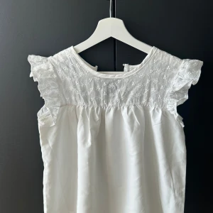 Vit blus med spets och volang - Supersöt vit blus med broderad spets upptill och volangdetaljer på axlarna. Blusen är ärmlös och har en luftig passform, perfekt för varma dagar. Blommigt broderimönster. 