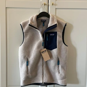 Fleeceväst från Patagonia - Säljer en beige fleeceväst från Patagonia med marinblå detaljer och dragkedjor. Västen har hög krage men kan vikas ner som enligt mig blir mycket snyggare, Perfekt nu för sommaren! Helt oanvänd och ny!😁