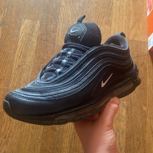 Nike Air Max 97 helsvarta sneakers - Innehåller inte lådan. Säljer ett par helsvarta Nike Air Max 97 sneakers med ikonisk vågig design och synlig luftdämpning i sulan. Skorna har snörning och en rund tå, samt diskret vit Nike-logga på sidan. Perfekta för dig som gillar klassiska och stilrena sneakers. 02