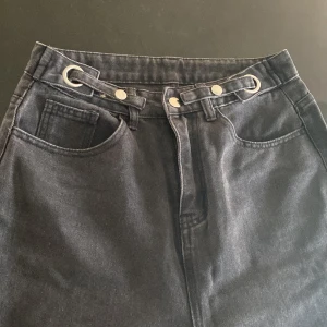 Gråa wide leg jeans med bältdetalj - Gråa jeans med hög midja och breda ben. Byxorna har en unik bältdetalj med metallringar och knappar framtill. Klassisk femficksmodell och raka ben. Innerbenslängd: 72 cm. Säljer då de är för korta för mig.
