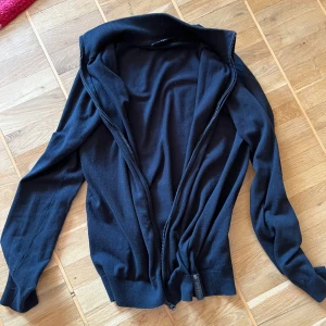 Svart stickad kofta med dragkedja - Svart stickad kofta med hög krage och hel dragkedja framtill. Tröjan har långa ärmar och ribbade muddar vid ärmslut och nederkant. Perfekt för lager-på-lager och enkel att matcha med olika outfits.