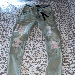  jeans från Amiri grå-rosa - Säljer ett par HELT NYA OANVÄNDA blågråa jeans från Amiri med tydliga slitningar och tryckt text på benen. Jeansen har klassisk femficksdesign, raka ben och detaljerade slitningar framtill. Perfekt för dig som gillar streetwear och vill sticka ut.